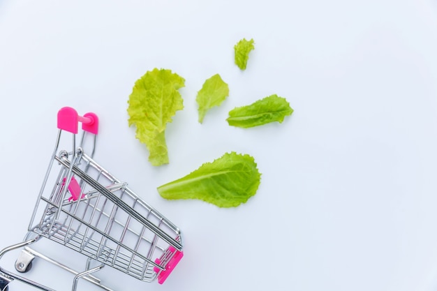 Kleiner Supermarktlebensmittel-Schubkarren zum Einkaufen mit grünen Salatblättern lokalisiert auf weißem Hintergrund