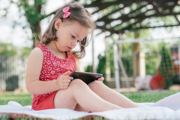 Kleines 3-4 Mädchen in roter Kleidung sitzt auf einer Decke auf grünem Gras und schaut ins Handy. Kinder, die Gadgets verwenden