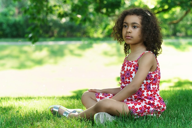 Kleines afrikanisches Mädchen, das auf einem Gras in einem Stadtpark sitzt