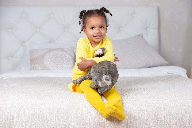 Kleines afroamerikanisches Mädchen in einem gelben Kleid sitzt mit einem weichen Spielzeug auf dem Bett.