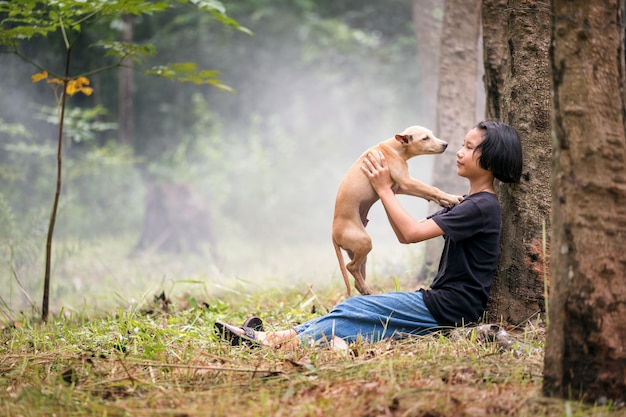 Kleines asiatisches Mädchen, das allein auf grünem Feld unter dem Baum mit ihrem Hund, im Freien an der Landschaft von Thailand sitzt