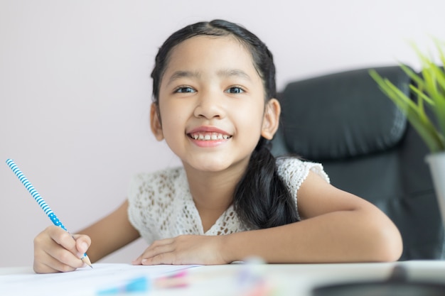 Kleines asiatisches Mädchen, das den Bleistift benutzt, um auf das Papier zu schreiben, das Hausaufgaben macht