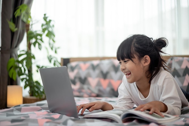 Kleines asiatisches Mädchen, das einen Laptop benutzt, während es auf dem Bett liegt