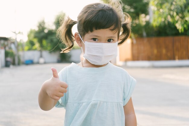 Kleines asiatisches Mädchen, das Maske zum Schutz pm2.5 trägt und Daumen hoch Geste für gute Luft im Freien zeigt. Luftverschmutzung pm2.5 und Coronavirus-Konzept