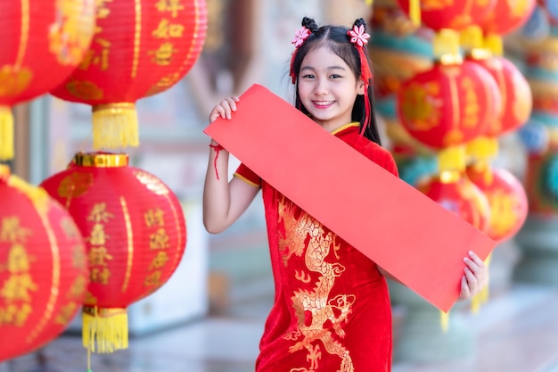 Kleines asiatisches Mädchen, das rote traditionelle chinesische Cheongsam-Dekoration trägt, zeigt leeres Papier rot und Laternen mit dem darauf geschriebenen chinesischen Text Segen ist ein Glückssegen für das chinesische Neujahr