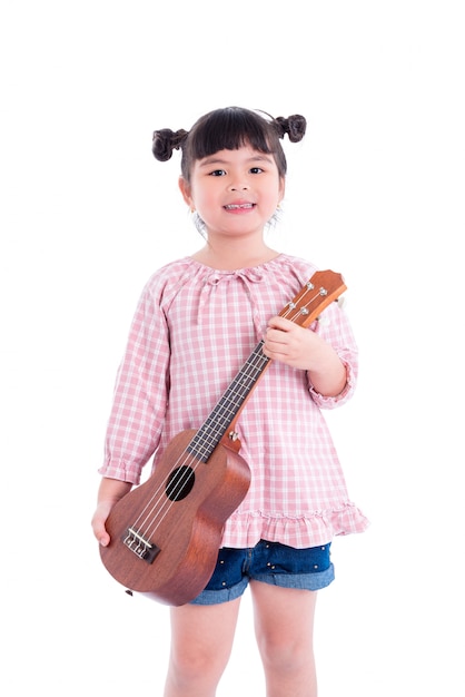 Kleines asiatisches Mädchen, das Ukulele und Lächeln über weißem Hintergrund hält