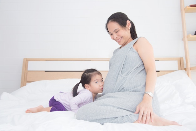 Kleines asiatisches Mädchen, das zu Hause ihren schwangeren Mutterbauch auf Bett küsst
