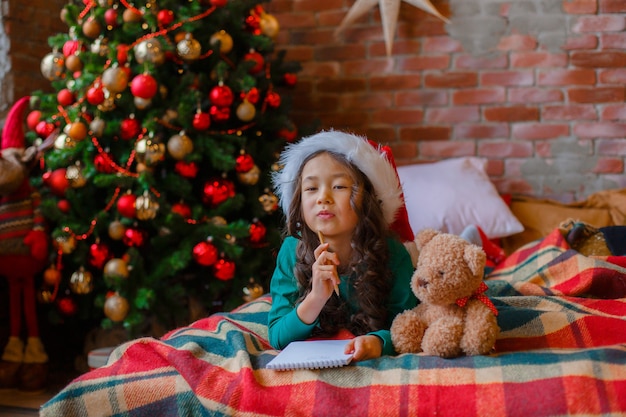 Kleines asiatisches Mädchen im Pyjama in der Nähe von Weihnachtsbaum schreibt einen Brief an den Weihnachtsmann Weihnachten Neujahr