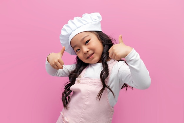 Kleines asiatisches Mädchen in Kochuniform lächelt und zeigt Vorlieben mit den Händen vor rosa isoliertem Hintergrund