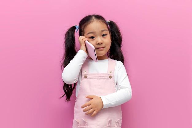 Kleines asiatisches Mädchen in rosafarbenem Sommerkleid, das auf rosa, isoliertem Hintergrund telefoniert