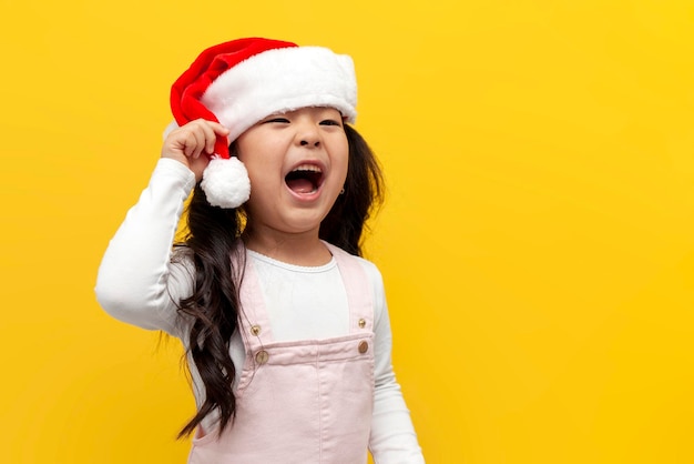 Kleines asiatisches Mädchen mit Weihnachtsmütze schreit laut auf gelbem Hintergrund, koreanisches Kind