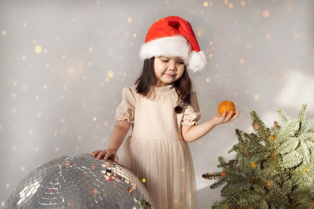 Kleines asiatisches Mädchen steht in Weihnachtsmütze in der Nähe der Discokugel und der Fichtenzweige. Hält eine Mandarine