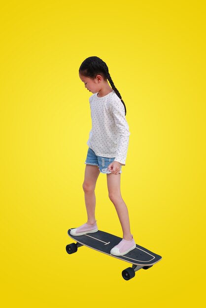 Kleines asiatisches Mädchenkind, das auf einem Skateboard lokalisiert auf gelbem Hintergrund eisläuft. Kind reitet auf Skateboard mit Beschneidungspfad