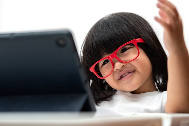 Kleines asiatisches Vorschulmädchen, das rote Brille trägt und Tablet-PC auf weißem Hintergrund verwendet, asiatisches Mädchen, das mit einem Videoanruf mit einem Tablet spricht und lernt, Bildungskonzept für Schulkinder.