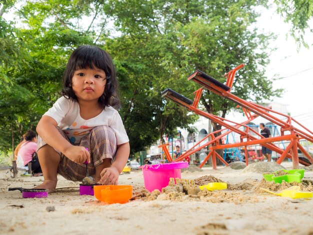 Kleines Asien-Mädchen, das im Sandkasten sitzt und mit Spielzeugsand-Schaufeleimer spielt