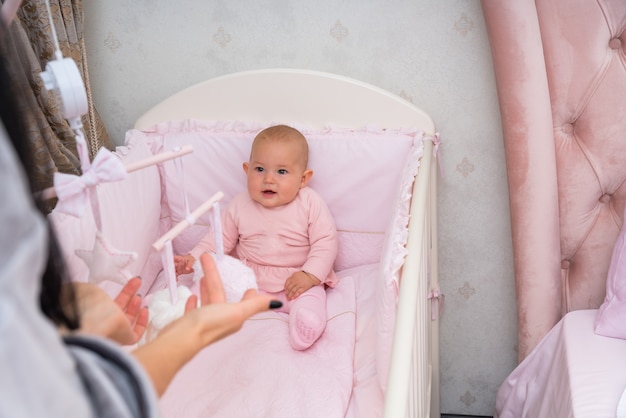 Kleines Baby beobachtet ein hängendes mobiles Spielzeug über ihrem Kinderbett, während ihre Mutter sich vorbeugt, um ihre Aufmerksamkeit zu erregen