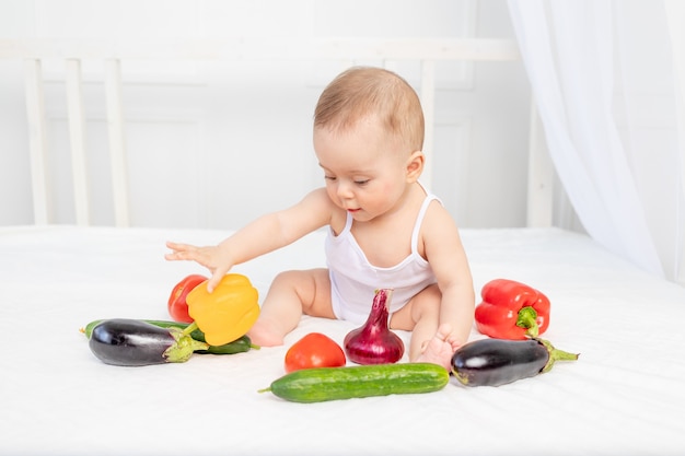 Kleines Baby, das auf dem Bett im Kinderzimmer mit Gemüse, Babynahrungskonzept sitzt,