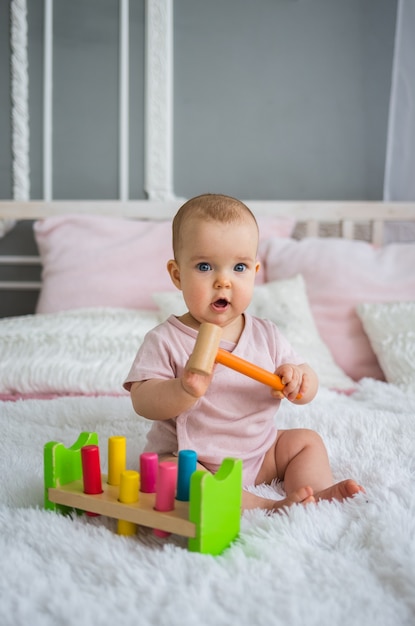 Kleines Baby in einem rosa Bodysuit sitzt mit einem hölzernen Lernspielzeug auf dem Bett im Raum