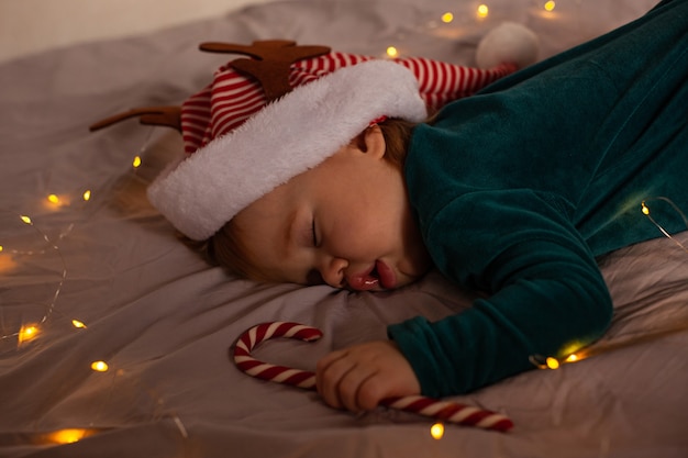 Kleines Baby liegt in einem Weihnachtskostüm und einer Mütze mit Hirschgeweih auf dem Bett. Weihnachten und Neujahr.