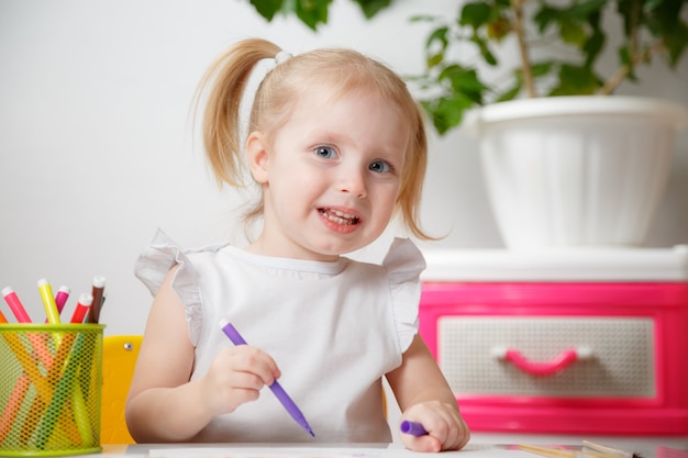 Kleines Baby malt am Tisch im Haus oder im Kindergarten Vorschule. Nettes entzückendes kleines Kind mit zwei Pferdeschwänzen, die Indoor.Box mit mehrfarbigen Markierungsstiften zeichnen.