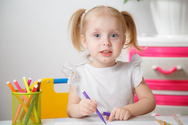 Kleines Baby malt am Tisch im Haus oder im Kindergarten Vorschule. Nettes entzückendes kleines Kind mit zwei Pferdeschwänzen, die Indoor.Box mit mehrfarbigen Markierungsstiften zeichnen.