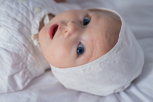 Kleines Baby mit blauen Augen im weißen Kleid und im Hut auf weißer Decke.