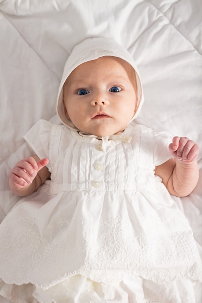 Kleines Baby mit blauen Augen im weißen Kleid und im Hut auf weißer Decke.