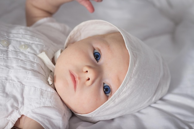 Kleines Baby mit blauen Augen im weißen Kleid und Mütze im Bett.