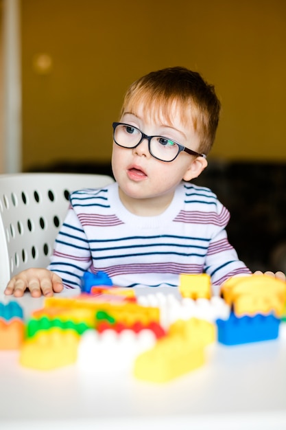 Kleines Baby mit Down-Syndrom mit den großen blauen Gläsern, die mit bunten Ziegelsteinen spielen