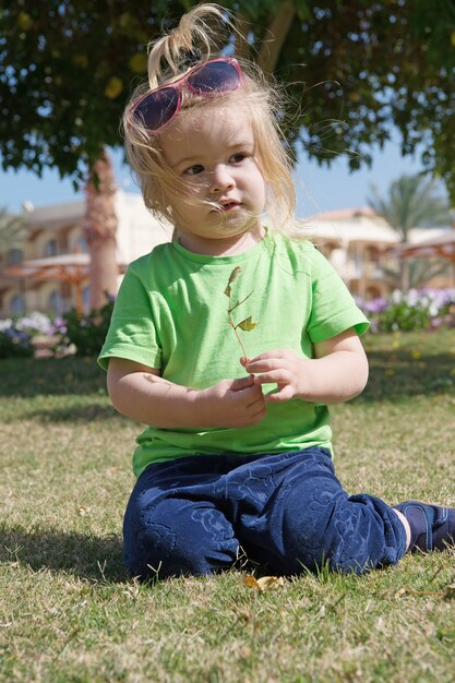 Kleines Baby oder süßes Kind mit entzückendem Gesicht und blonden Haaren in Hemd, Sonnenbrille und Hose, das auf grünem Gras sitzt und mit Blättern im Freien auf natürlichem Hintergrund spielt