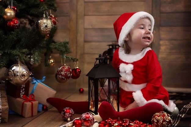 Kleines Baby sitzt auf dem Boden im Zimmer vor einem geschmückten Weihnachtsbaum