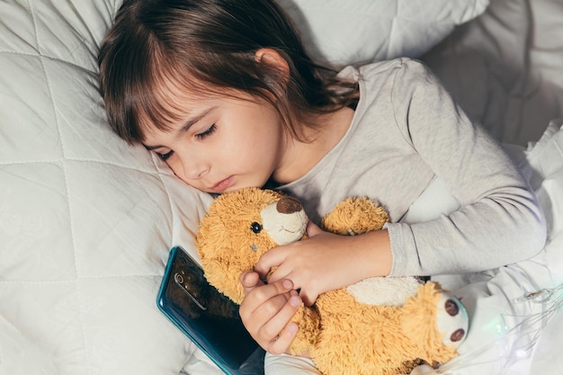 Kleines Babymädchen, das im Bett mit Telefon und ihrem weichen Teddybärspielzeug schläft