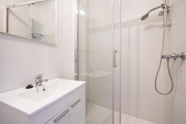 Kleines Badezimmer mit weiß lackierten Holzschränken mit rahmenlosen Türen, Spiegel an der Wand, Edelstahlarmaturen, Porzellanwaschbecken und ebenerdiger Dusche mit Glastrennwänden