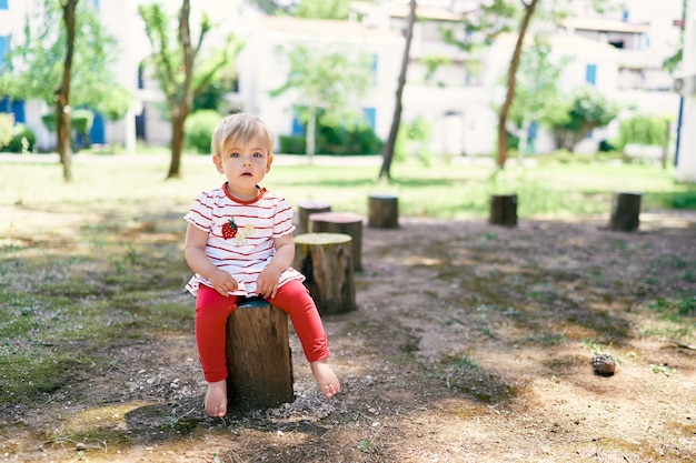 Kleines barfüßiges Baby sitzt auf einem Baumstumpf im Hof