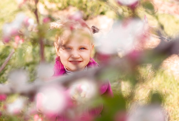 Kleines blauäugiges blondes Mädchen in Rosa schaut hinter Ästen eines blühenden Baumes in die Kamera