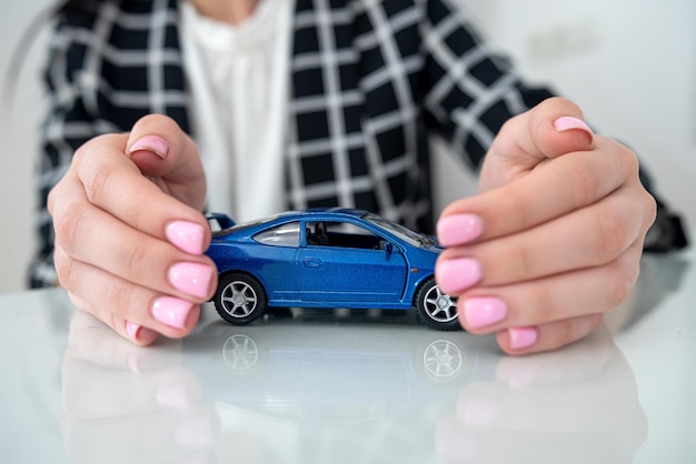 Kleines blaues Spielzeugauto und eine weibliche Hand am Schreibtisch als Versicherung Schutz des Automobilkonzepts