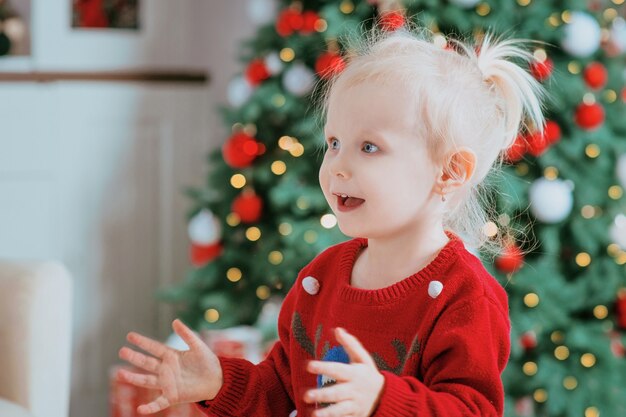 kleines blondes kaukasisches Mädchen im roten Pullover mit Seifenblasen am Weihnachtsbaum