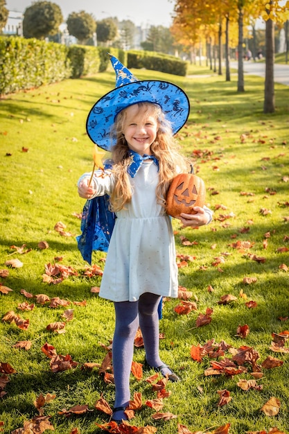 Kleines blondes Mädchen, das als Hexenzauberer für Halloween-Hutmantel, Kürbislachen, Lächeln, Herbstferien gekleidet ist