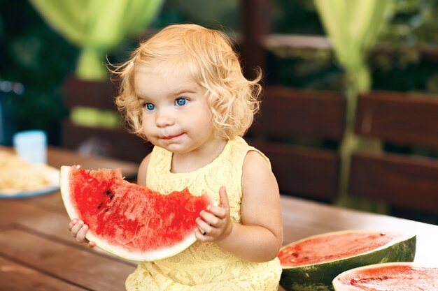 Kleines blondes Mädchen, das eine saftige Wassermelone im Garten isst. Kinder essen Obst auf der Straße. Gesundes Essen für Kinder. Gartenarbeit für Kleinkinder.