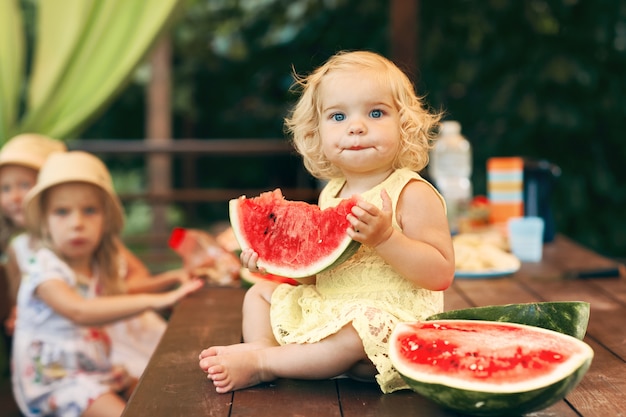 Kleines blondes Mädchen, das eine saftige Wassermelone im Garten isst. Kinder essen Obst auf der Straße. Gesundes Essen für Kinder. Gartenarbeit für Kleinkinder.