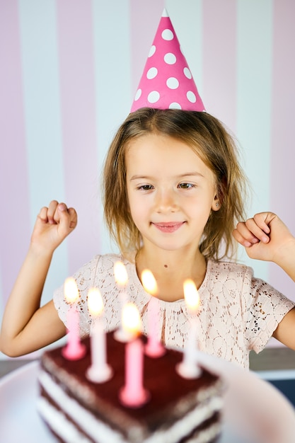 Kleines blondes Mädchen, das im Geburtstag rosa Kappe, eine Schokoladengeburtstagstorte mit Kerzen lächelt. Kind feiert ihren Geburtstag. Alles Gute zum Geburtstag.