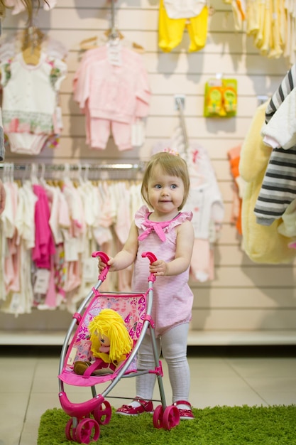 Kleines blondes Mädchen, das im Kinderladen mit dem Spielzeugkinderwagen lächelt und in die Hände klatscht, Nahaufnahme