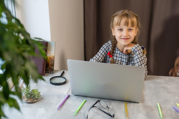 Kleines blondes Mädchen, das zu Hause am Tisch Hausaufgaben macht. Das Kind wird zu Hause unterrichtet. Ein Mädchen mit hellem Haar führt eine Aufgabe online mit einem Laptop und einem Tablet-Computer aus.