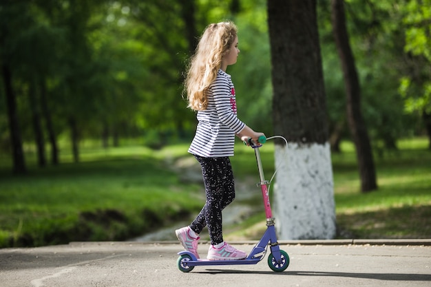 Kleines blondes Mädchen fährt mit dem Roller im Park.