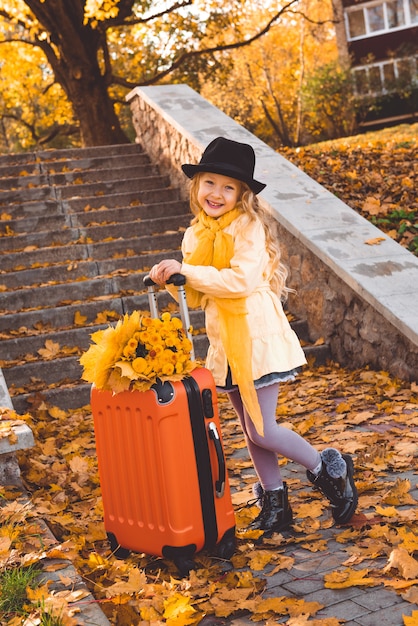 Kleines blondes Mädchen im Herbst mit Blumen und Koffer