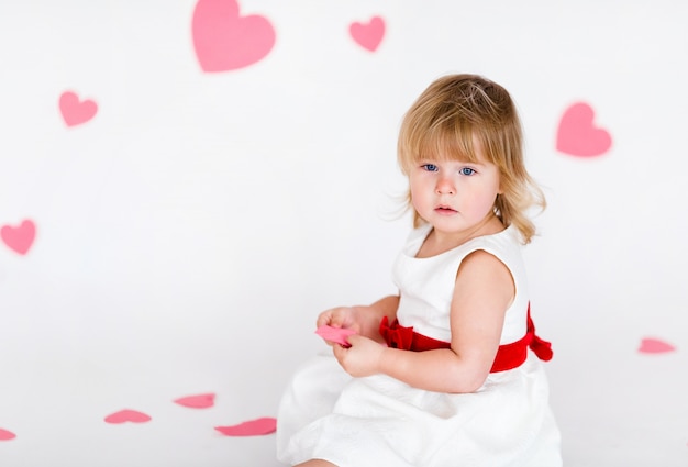 Kleines blondes Mädchen im weißen Kleid mit rotem Band auf weißem Boden mit rosa Herzen am Valentinsgrußtag