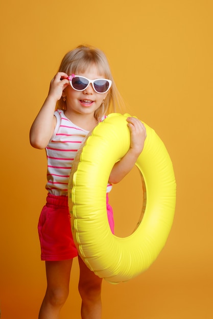 kleines blondes Mädchen in der Sommerkleidung, die Sonnenbrille mit einem Schwimmkreis auf gelbem Hintergrund trägt. Das Konzept der Sommerferien