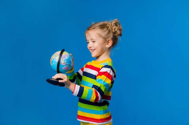 Kleines blondes Mädchen in einem gestreiften Pullover hält einen Globus auf blauem Hintergrund