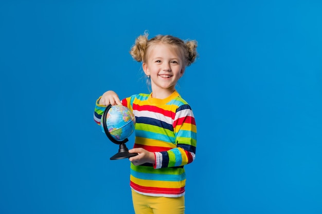 Kleines blondes Mädchen in einem gestreiften Pullover hält einen Globus auf blauem Hintergrund