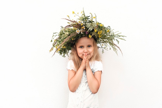 Kleines blondes Mädchen in einem Kranz von Wildblumen auf einem weißen Wandhintergrund